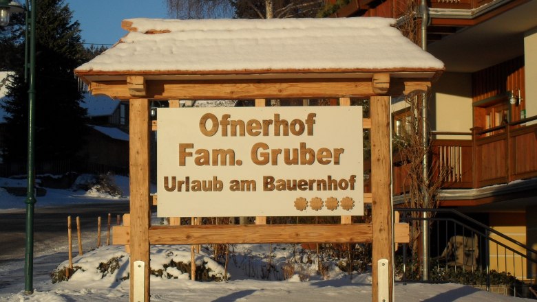 Holzschild mit der Aufschrift 'Ofnerhof Fam. Gruber Urlaub am Bauernhof' im Schnee.