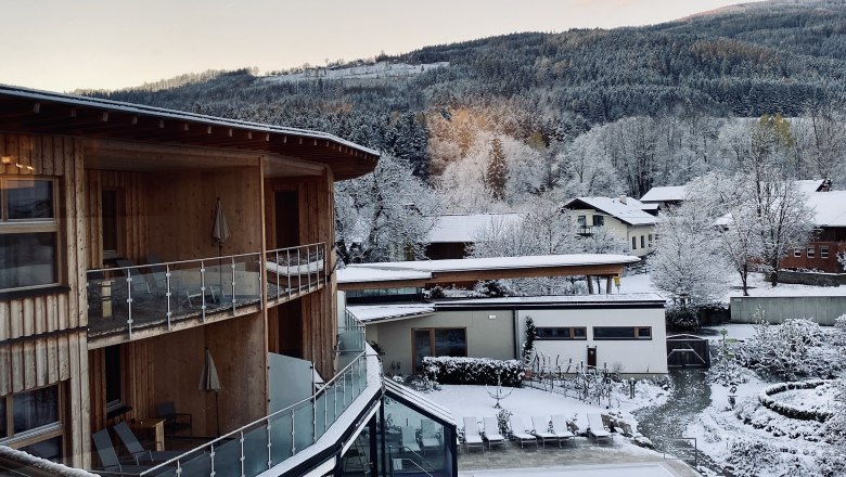 Winterliche Landschaft mit schneebedeckten Gebäuden und Bäumen, im Vordergrund ein modernes Holzgebäude mit Balkonen.