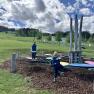 Zwei Kinder spielen auf einem Kletterger&uuml;st in einer gr&uuml;nen Landschaft mit B&auml;umen und Wolken am Himmel.