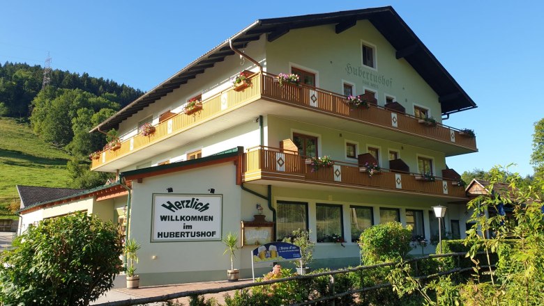 Ein traditionelles Gasthaus namens Hubertushof mit Blumen auf den Balkonen, umgeben von grüner Landschaft und blauem Himmel.