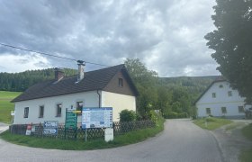 Ländliche Straße mit zwei Häusern und Wegweisern, umgeben von Wäldern und Wiesen.