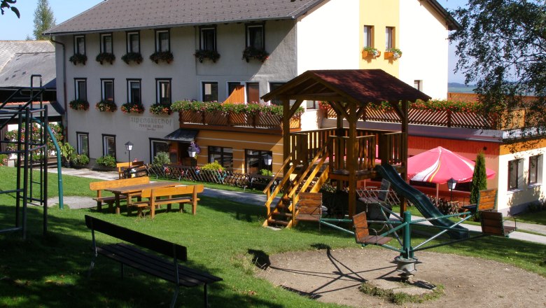 Ein Gasthof mit Spielplatz im Vordergrund, umgeben von gr&uuml;ner Wiese und B&auml;umen.