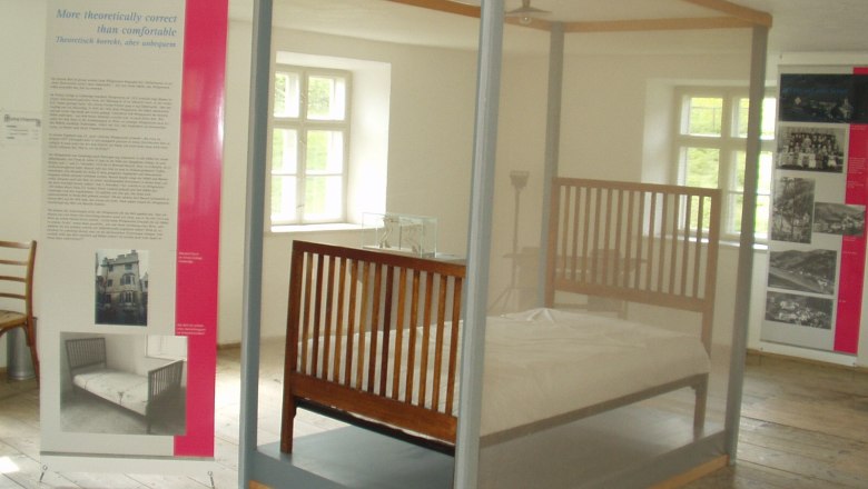 Ausstellungsraum mit einem Bett hinter Glas und Informationstafeln.