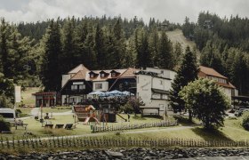 Ein Gasthaus in einer bergigen Landschaft mit Spielplatz und Wald im Hintergrund.