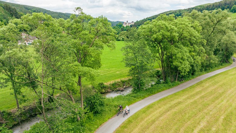 Zwei Radfahrer auf einem Weg entlang eines Flusses, umgeben von gr&uuml;nen B&auml;umen und Wiesen.