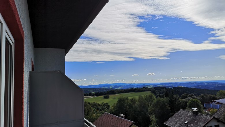 Blick von einem Balkon auf eine Landschaft mit H&auml;usern, B&auml;umen und H&uuml;geln unter einem blauen Himmel mit Wolken.