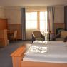 Ein gemütliches Hotelzimmer mit Doppelbett, Schreibtisch, Fernseher und zwei Sesseln vor einem Fenster mit Vorhängen.