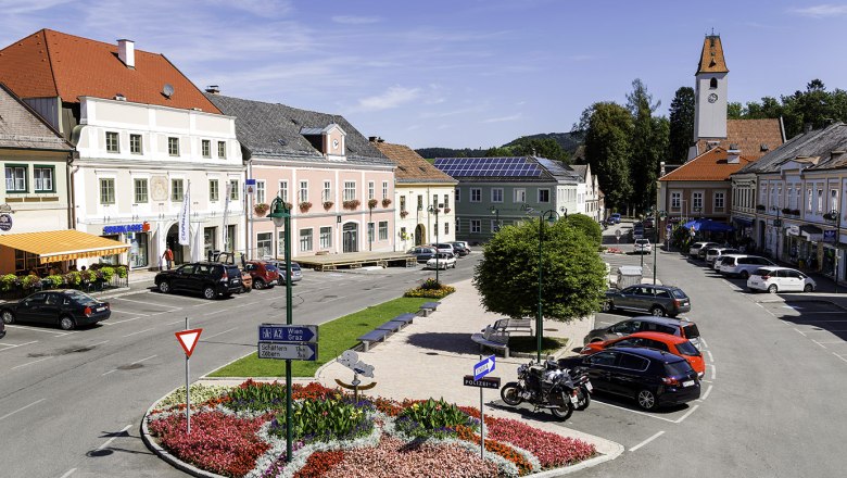 Hauptplatz in Aspang Markt mit bunten Blumenbeeten, geparkten Autos und historischen Geb&auml;uden.
