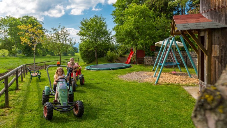 Zwei Kinder fahren auf Spielzeugtraktoren &uuml;ber eine Wiese. Im Hintergrund sind eine Schaukel, eine Rutsche und ein Trampolin zu sehen.