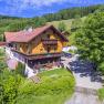 Ein traditionelles Holzhaus in einer gr&uuml;nen, h&uuml;geligen Landschaft mit B&auml;umen und einem blauen Himmel.