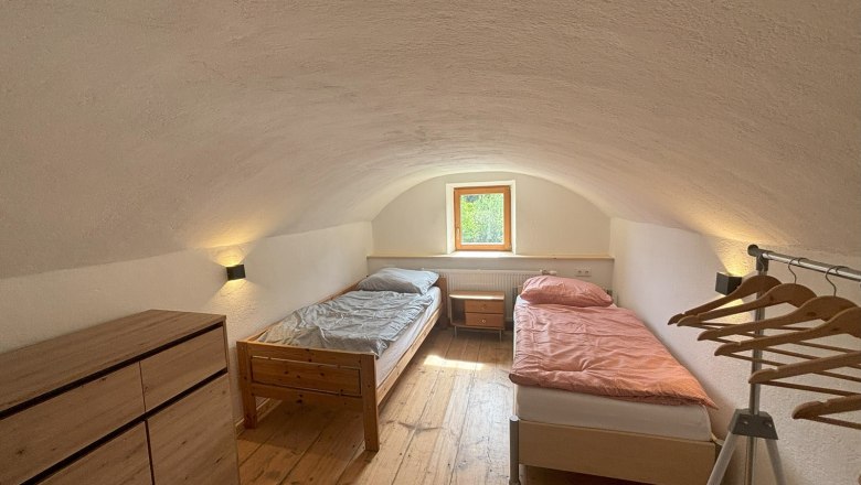 Ein einfaches Doppelzimmer mit zwei Einzelbetten, einer Kommode und einem Kleiderst&auml;nder. Ein kleines Fenster l&auml;sst Tageslicht herein.