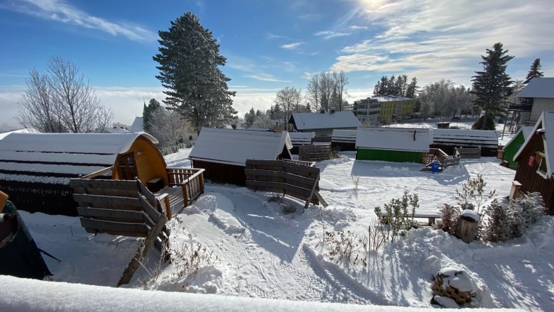 Winterlicher Glamping-Park mit schneebedeckten Hütten und Bäumen unter blauem Himmel und Sonnenschein.
