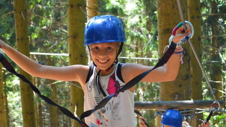 Ein Mädchen mit blauem Helm balanciert auf einem Kletterparcours im Wald.