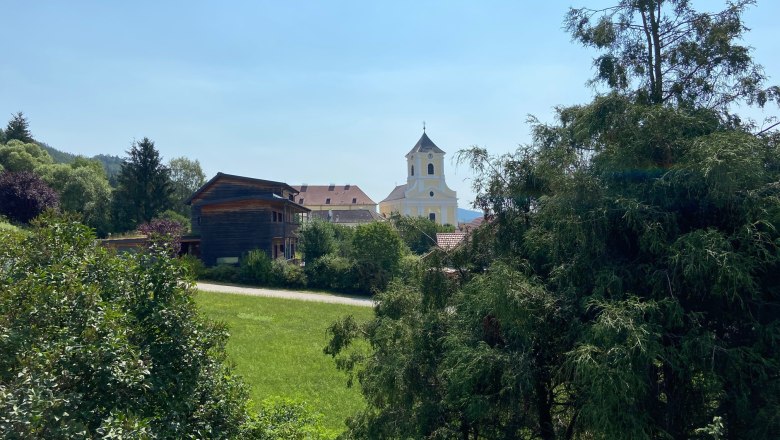 Blick auf eine Kirche und ein Holzhaus hinter B&auml;umen und Wiesen.