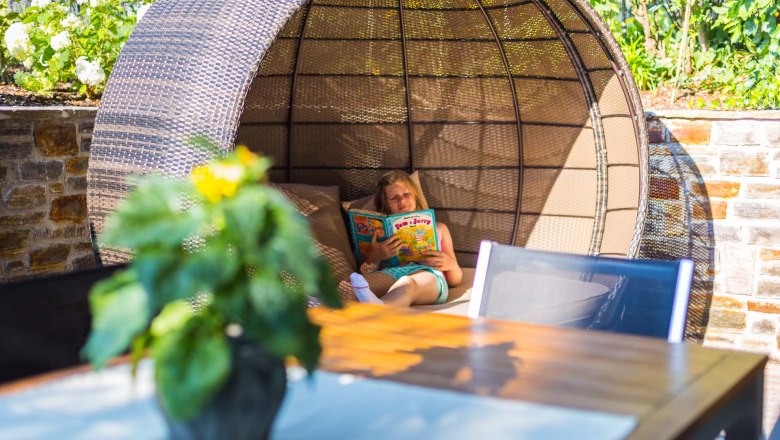 Person liest ein Buch in einer runden, geflochtenen Sitzgelegenheit im Freien.