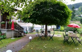 Ein gemütlicher Gastgarten mit Tischen und Stühlen unter einem großen Baum, umgeben von grüner Landschaft.