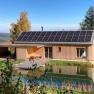Ein modernes Holzhaus mit Solarpanelen auf dem Dach, umgeben von Natur und einem kleinen Teich im Vordergrund.