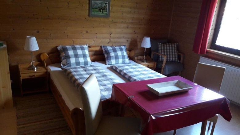 Gemütliches Zimmer mit Holzwänden, Doppelbett, Tisch mit roter Tischdecke und Stühlen, Lampe und Fenster mit rotem Vorhang.