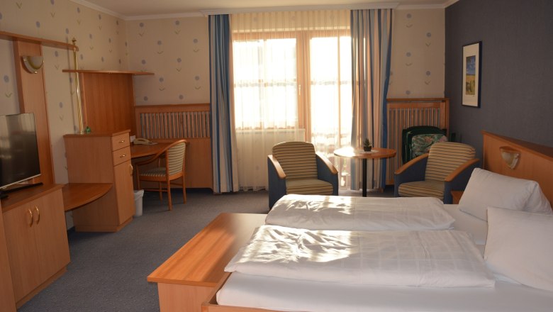 Ein gemütliches Hotelzimmer mit Doppelbett, Schreibtisch, Fernseher und zwei Sesseln vor einem Fenster mit Vorhängen.