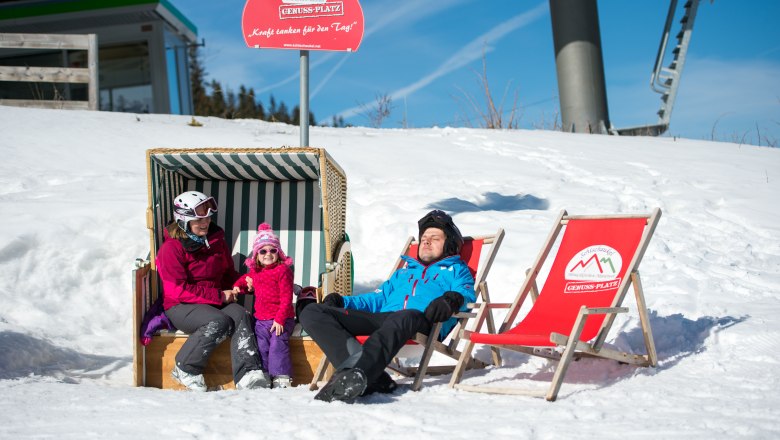 Familie entspannt im Schnee auf der Erlebnisalm, sitzend in einem Strandkorb und Liegestühlen unter einem Schild mit der Aufschrift 'Genussplatz'.