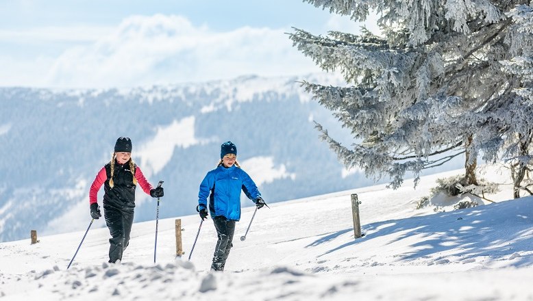 Zwei Personen beim Langlaufen auf einer verschneiten Loipe mit Bergen im Hintergrund.