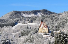 Winterlandschaft mit Kirche vor bewaldetem H&uuml;gel.